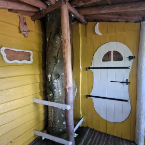 Cabane en bois colorée avec une porte blanche en forme de visage et un arbre à l'intérieur.