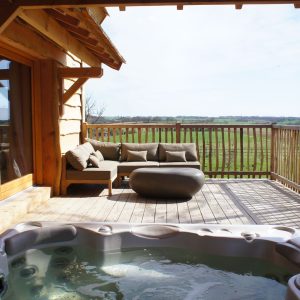 Cabane en bois avec jacuzzi sur terrasse, vue dégagée sur la campagne.