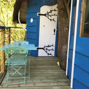 Cabane perchée bleue avec une porte originale et une terrasse en bois, entourée d'arbres.