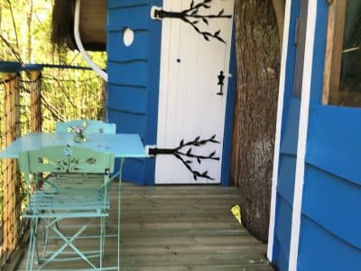 Cabane perchée bleue avec une porte originale et une terrasse en bois, entourée d'arbres.