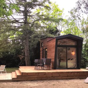 Cabane moderne en bois avec terrasse en bois, entourée d'arbres verdoyants et mobilier extérieur.