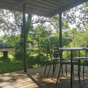 Terrasse en bois d'un chalet au bord de l'eau, entourée d'arbres verdoyants.