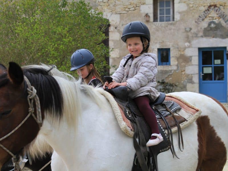 Séjour à la ferme équestre : enfants souriants à cheval devant une belle bâtisse.