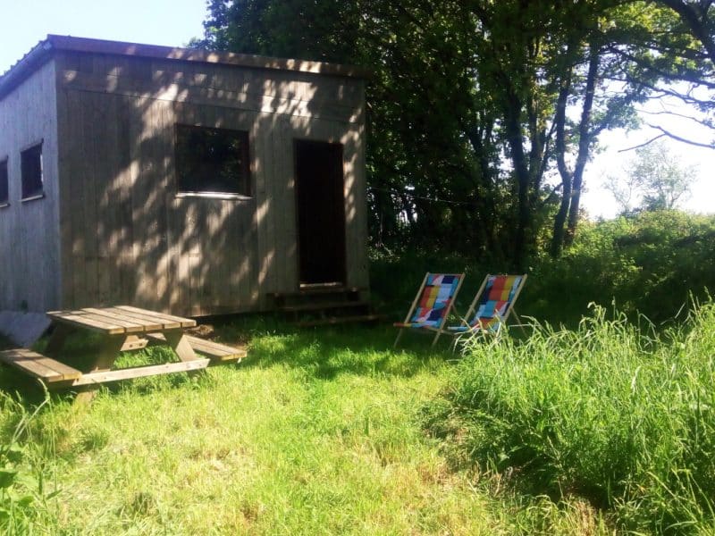 Cabane en bois entourée de verdure, avec une table en bois et des tableaux colorés.
