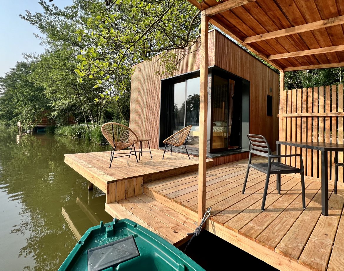 Cabane flottante en bois avec terrasse sur pilotis, vue sur l'eau et verdure environnante.