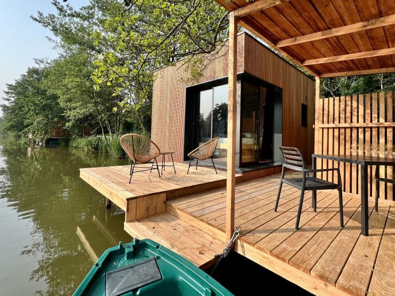 Cabane flottante en bois avec terrasse sur pilotis, vue sur l'eau et verdure environnante.