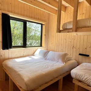 Cabane en bois avec lits superposés, lumière naturelle et vue sur la verdure.