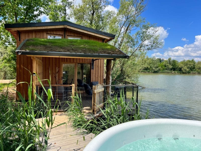 Cabane en bois au bord de l'eau, avec un toit végétal et un jacuzzi sur la terrasse.