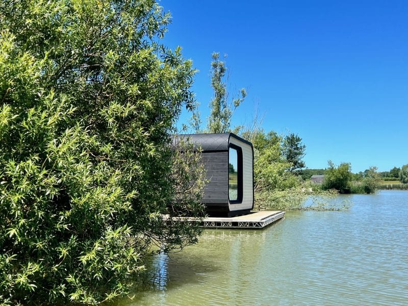 Cabane flottante moderne sur un lac, entourée de verdure et sous un ciel bleu.