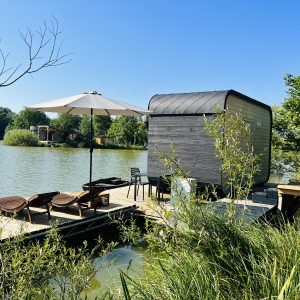 Cabane flottante moderne au bord d'un lac, avec parasol et chaises longues sur la terrasse.