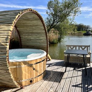 Hébergement insolite : un jacuzzi en bois sur une terrasse au bord de l'eau, entouré de verdure.