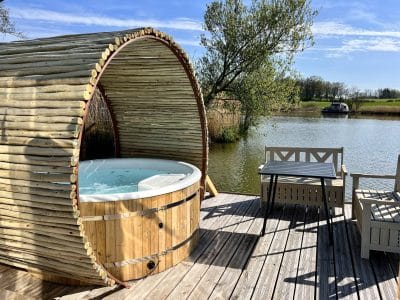 Hébergement insolite : un jacuzzi en bois sur une terrasse au bord de l'eau, entouré de verdure.