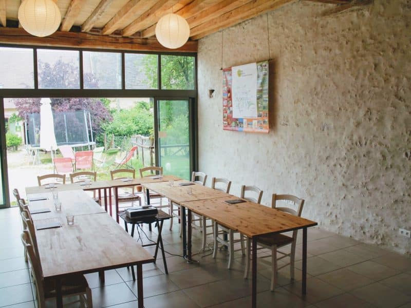 Salle de réunion dans un gîte, avec grandes fenêtres et tables en bois.