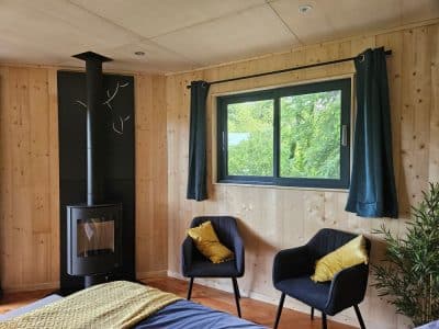 Chalet en bois avec poêle à bois, fenêtres donnant sur la verdure et fauteuils confortables.