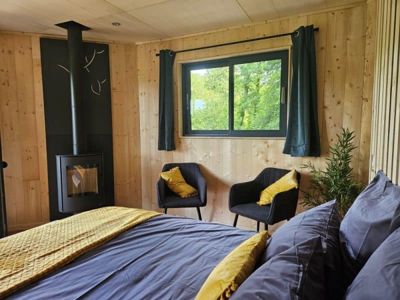 Chalet en bois avec un lit confortable, poêle à bois et vue sur la nature verdoyante.
