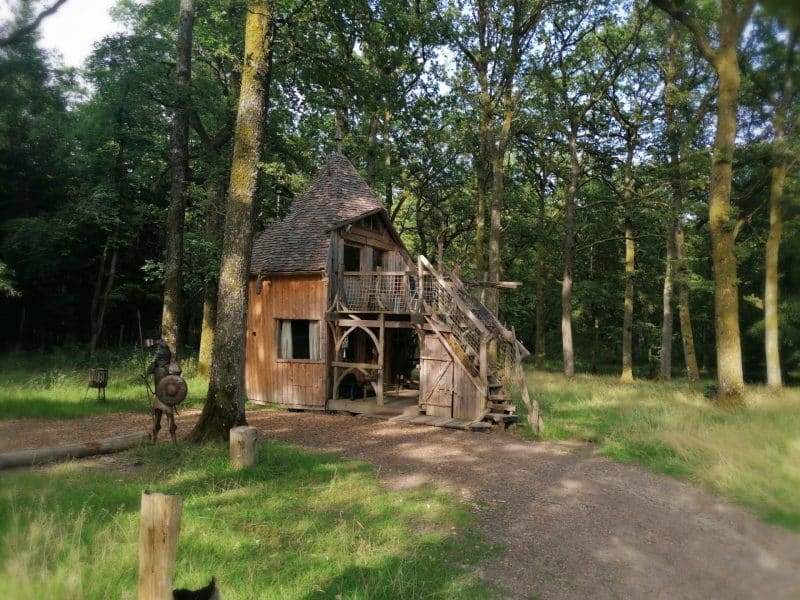 Cabane en bois perchée dans les arbres, entourée de verdure et de grands chênes.