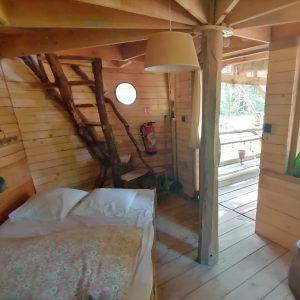 Cabane en bois avec un lit douillet, escaliers en rondins et lumière naturelle abondante.