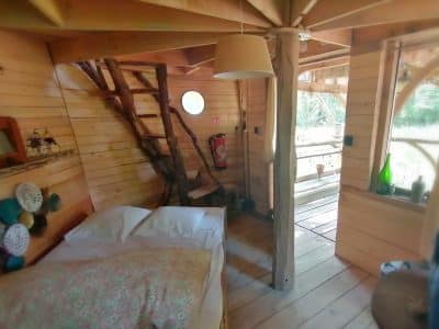 Cabane en bois avec un lit douillet, escaliers en rondins et lumière naturelle abondante.