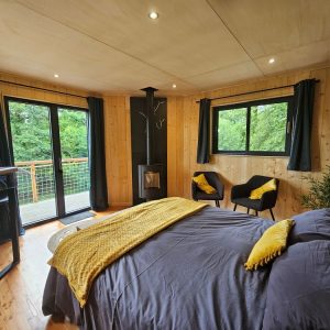 Chalet moderne avec grandes fenêtres, lit douillet et touches de jaune, entouré de verdure.