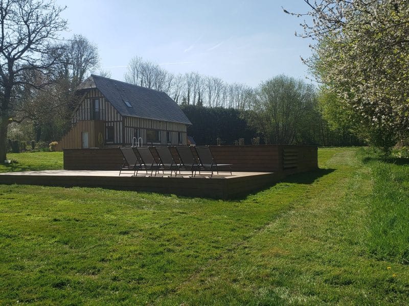 Charmante maison de campagne avec terrasse en bois et chaises longues, entourée de verdure.