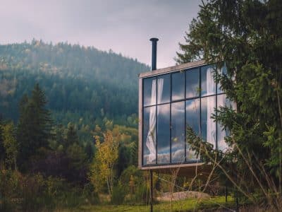 Cabane moderne en verre, entourée de verdure, reflétant les montagnes environnantes.