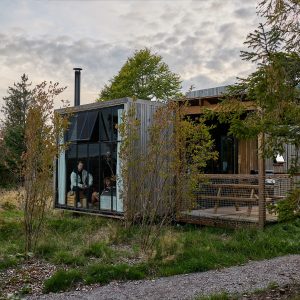 Cabane moderne en bois avec grandes fenêtres, entourée de verdure et d'un chemin en gravier.