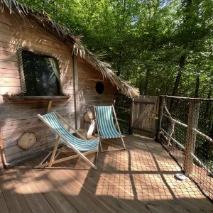 Cabane dans les arbres avec terrasse en bois, chaises longues et vue sur la forêt verdoyante.