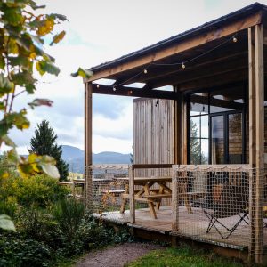 Cabane en bois avec terrasse, entourée de verdure et vue sur les montagnes. Ambiance chaleureuse.