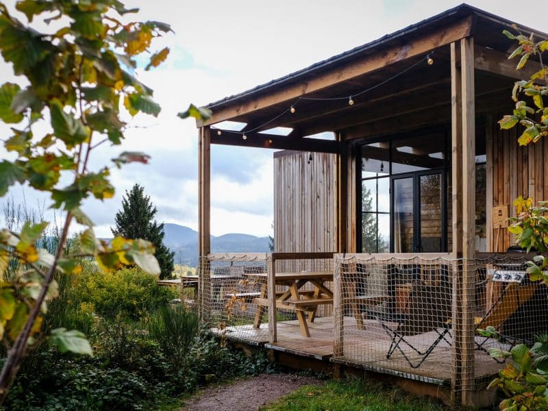 Cabane en bois avec terrasse, entourée de verdure et vue sur les montagnes. Ambiance chaleureuse.
