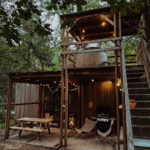 Cabane perchée en bois avec terrasse, éclairage chaleureux et coin barbecue en pleine nature.