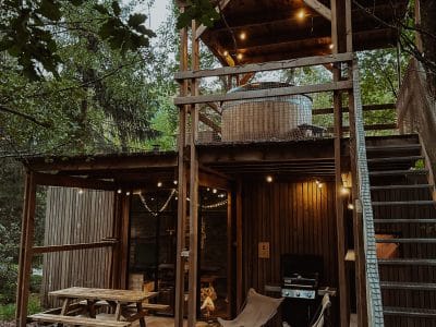Cabane perchée en bois avec terrasse, éclairage chaleureux et coin barbecue en pleine nature.