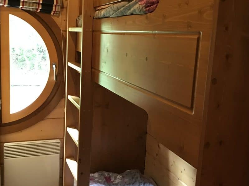 Cabane en bois avec lits superposés et fenêtre ronde, offrant une ambiance chaleureuse et cosy.