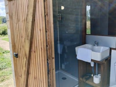 Cabane en bois avec salle de bain moderne, douche et lavabo, entourée de verdure.
