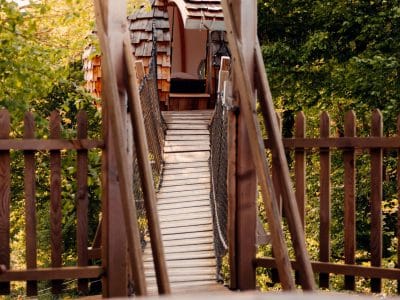 Cabane perchée accessible par un pont suspendu, entourée de verdure et d'une piscine.
