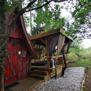 Cabane en bois rouge avec terrasse surélevée, entourée d'arbres verdoyants.
