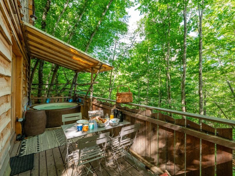Cabane dans les arbres avec terrasse, jacuzzi et vue sur la forêt verdoyante.