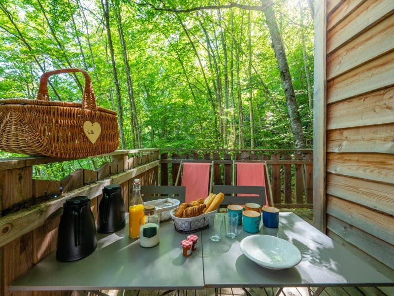 Cabane dans les arbres avec une terrasse, vue sur la forêt et un petit déjeuner appétissant.