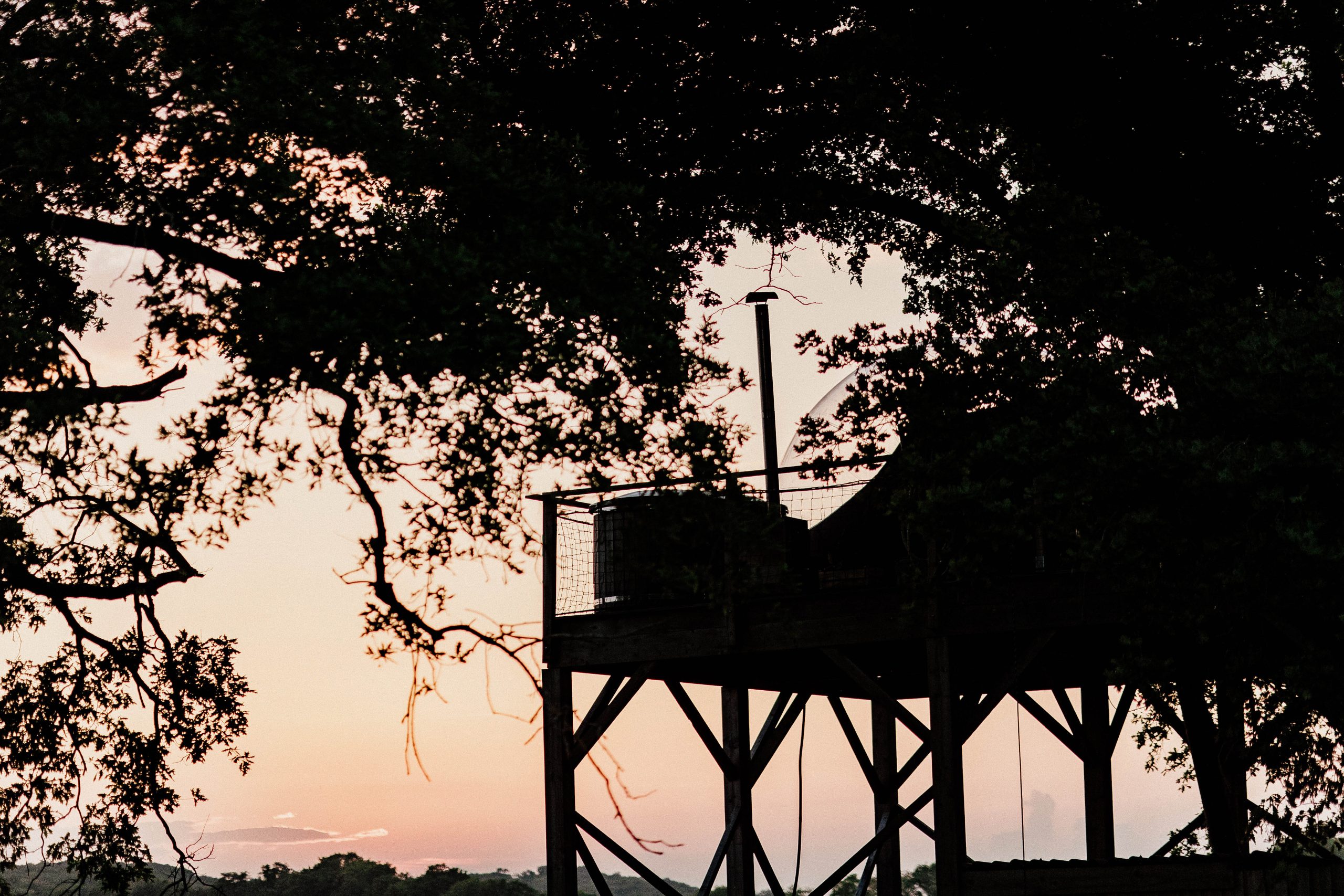 Cabane perchée sur pilotis, entourée d'arbres, au coucher de soleil. Ambiance paisible et naturelle.
