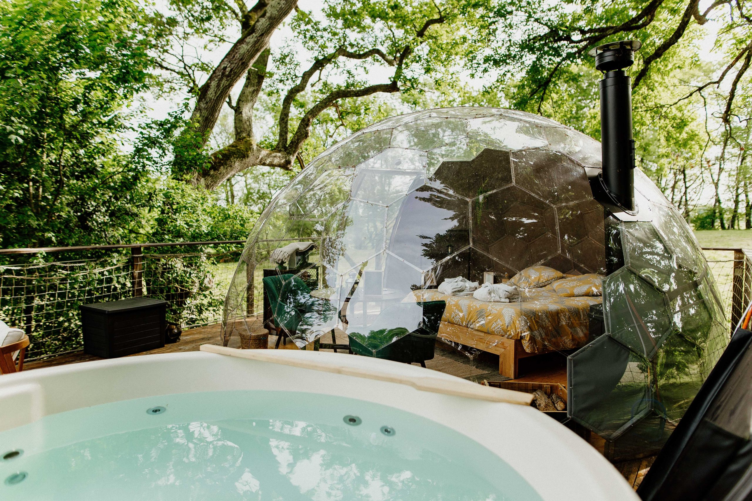 Hébergement insolite : bulle transparente avec lit douillet et jacuzzi, entourée de verdure.