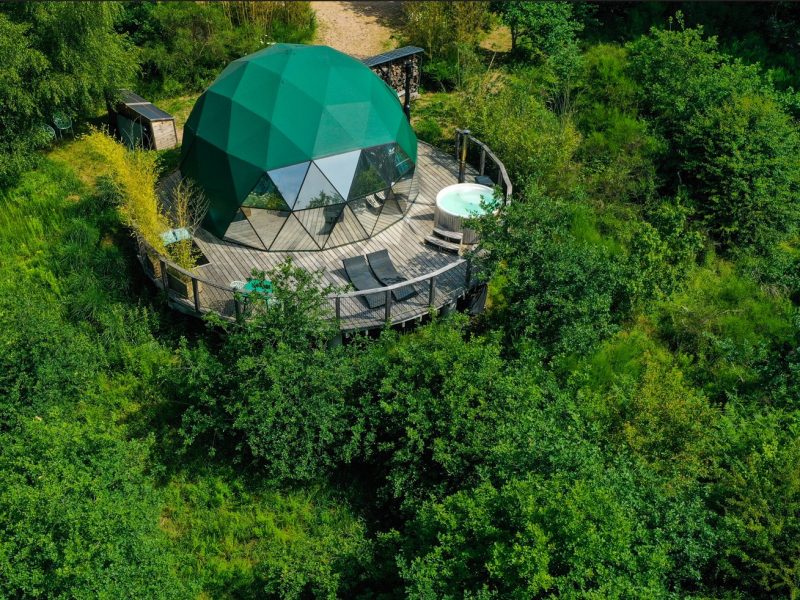 Dôme géodésique vert entouré de verdure, avec terrasse et jacuzzi en pleine nature.
