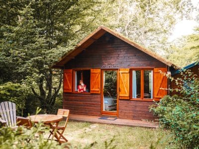 Charmant chalet en bois avec volets ouverts, entouré de verdure et d'un coin repas extérieur.