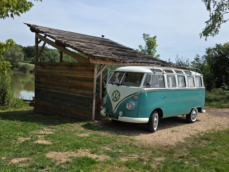 Hébergement insolite : un combi Volkswagen vintage sous un abri en bois, au bord de l'eau.
