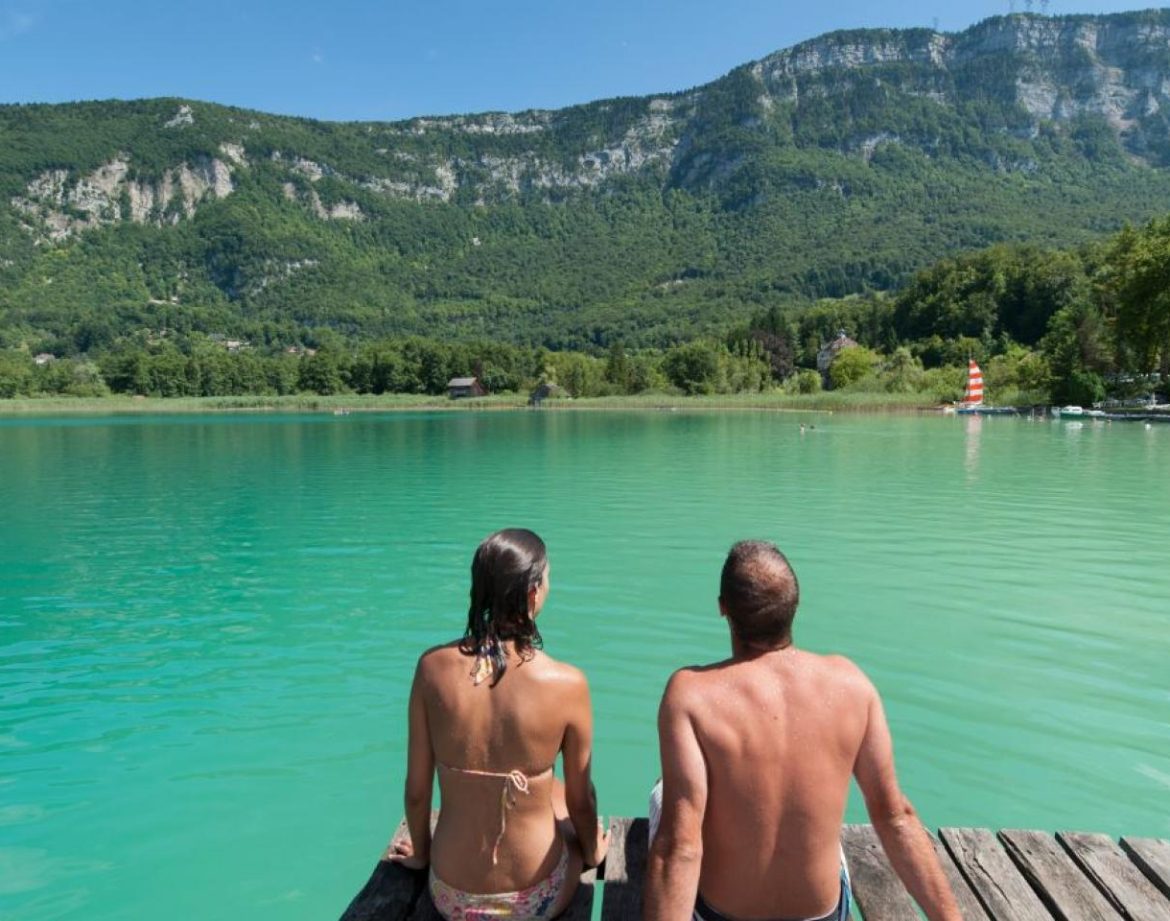 Un couple se détend sur un ponton en bois, face à un lac turquoise entouré de montagnes verdoyantes.