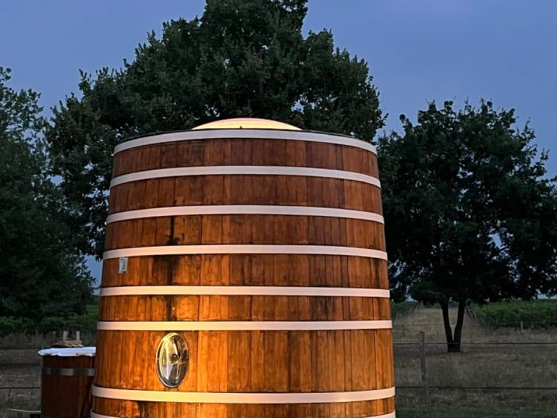 Hébergement insolite : un baril en bois éclairé, entouré d'arbres, sous un ciel nuageux.
