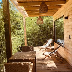 Cabane en bois avec terrasse ensoleillée, chaises longues et vue sur un jardin de bambous.