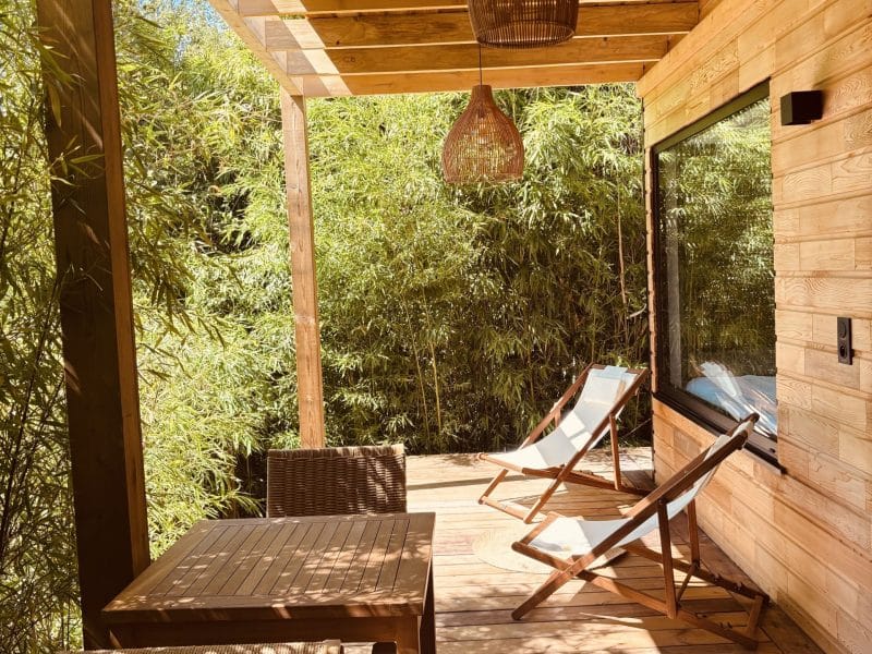 Cabane en bois avec terrasse, chaises longues et ambiance verdoyante.
