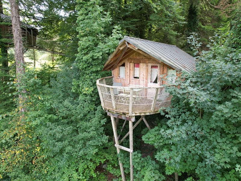 Cabane perchée dans les arbres, avec un balcon en bois entouré de verdure luxuriante.