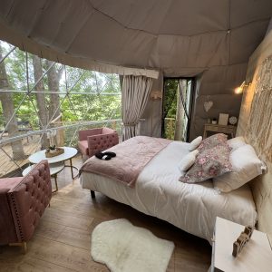 Dôme transparent en pleine nature, lit douillet et coin salon avec vue sur les arbres.