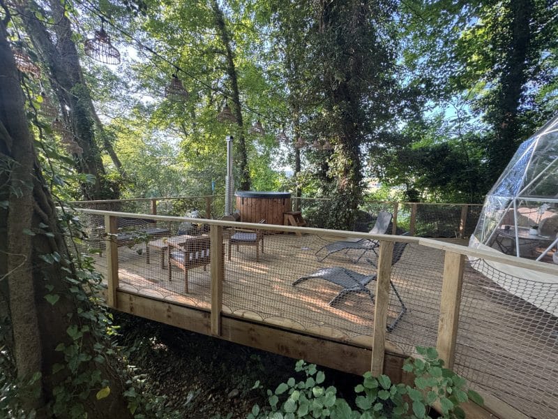 Cabane perchée dans les arbres avec terrasse en bois et jacuzzi, entourée de verdure.