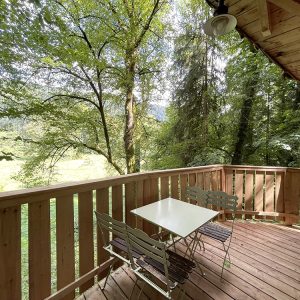 Cabane en bois avec terrasse ensoleillée, entourée d'une forêt verdoyante.